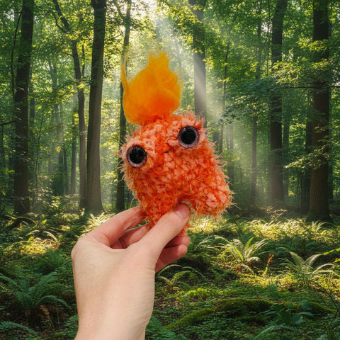 Hand holding a fluffy orange creature toy with a yellow top against a green park background