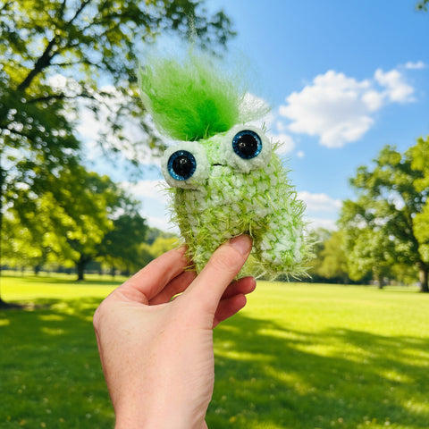 Hand holding a green plush toy with black eyes against a neutral background