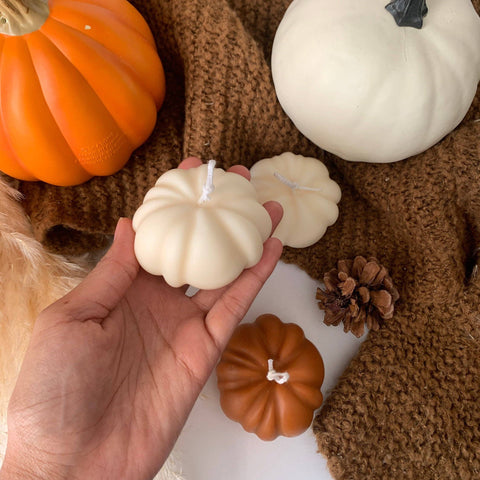 Hand holding a white pumpkin-shaped candle with orange and white pumpkins on a brown textured surface.