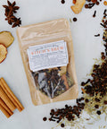 A kraft paper pouch labeled 'Witch's Brew' containing herbal tea, styled in a flat lay with cinnamon sticks, star anise, dried apple slices, and scattered spices on a white marble background.