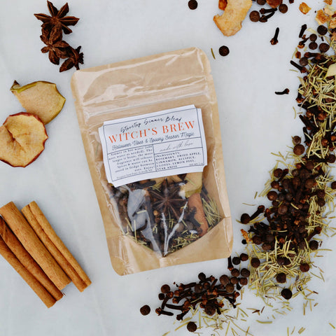 A kraft paper pouch labeled 'Witch's Brew' containing herbal tea, styled in a flat lay with cinnamon sticks, star anise, dried apple slices, and scattered spices on a white marble background.