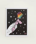 Framed artwork of a hand holding a rainbow rose against a starry night sky on a pink wall.