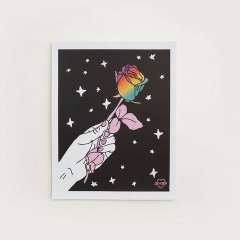 Framed artwork of a hand holding a rainbow rose against a starry night sky on a pink wall.