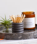 Dark grey colored match holder with matches on a marble tray with a plant and magazine underneath.