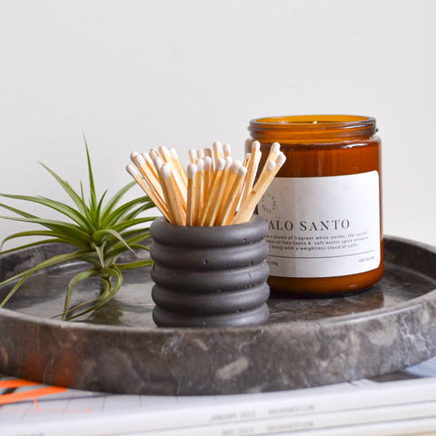 Dark grey colored match holder with matches on a marble tray with a plant and magazine underneath.