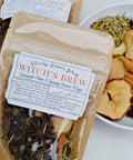 A kraft paper pouch labeled 'Witch's Brew', filled with dried herbs and spices, shown next to a white plate containing cinnamon sticks, dried apple slices, cloves, and other ingredients on a light surface.