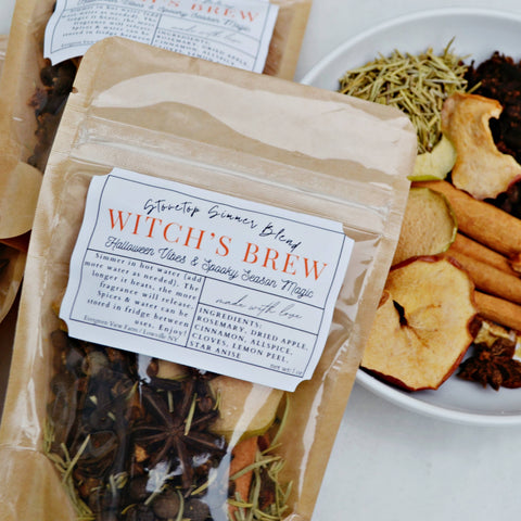 A kraft paper pouch labeled 'Witch's Brew', filled with dried herbs and spices, shown next to a white plate containing cinnamon sticks, dried apple slices, cloves, and other ingredients on a light surface.