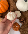 Hand holding a white pumpkin-shaped candle with orange and white pumpkins on a brown textured surface.
