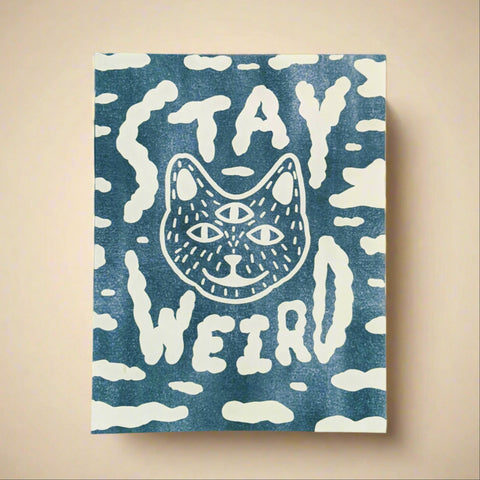 Blue towel with white cat illustration and 'Stay Weird' text on a blue and gray background