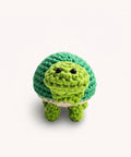 A green crocheted plush turtle toy with black bead eyes, standing on a clear glass cylinder against a plain light background.
