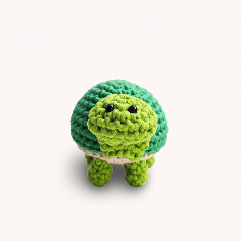A green crocheted plush turtle toy with black bead eyes, standing on a clear glass cylinder against a plain light background.