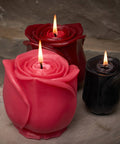 Three rose-shaped candles in pink, red, and black on a stone surface with a stone wall background.