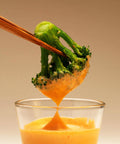 Broccoli being dipped into a glass of cheese sauce with wooden tongs.