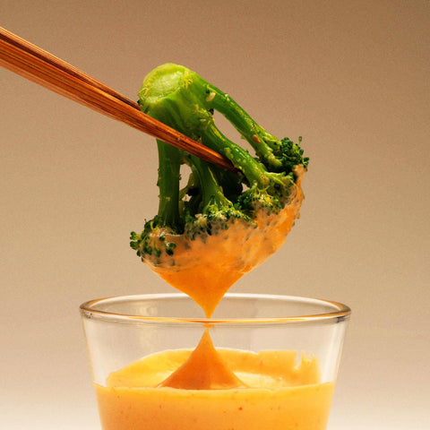 Broccoli being dipped into a glass of cheese sauce with wooden tongs.