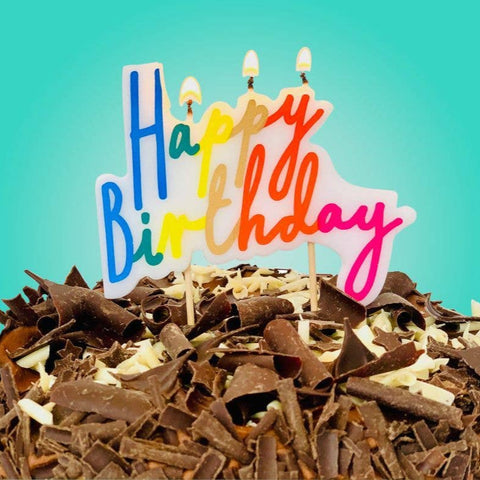 Chocolate birthday cake with colorful 'Happy Birthday' topper against a teal background