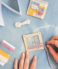 Person using a small loom with colorful threads on a light blue surface.