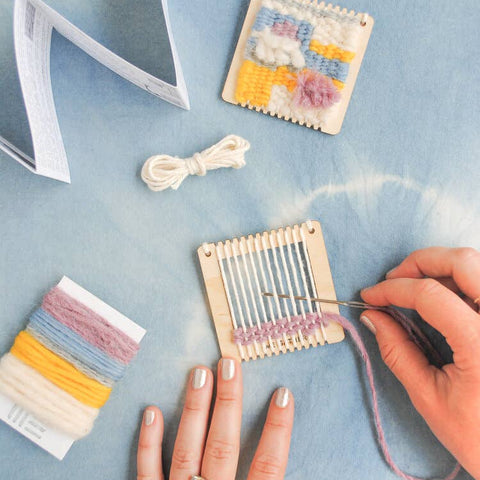Person using a small loom with colorful threads on a light blue surface.