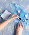 Person applying blue paint to denim fabric with various paint supplies on a blue surface.