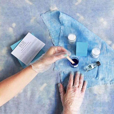 Person applying blue paint to denim fabric with various paint supplies on a blue surface.