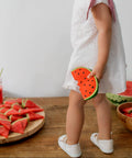Child in a white dress with watermelon decorations and slices on a wooden table.