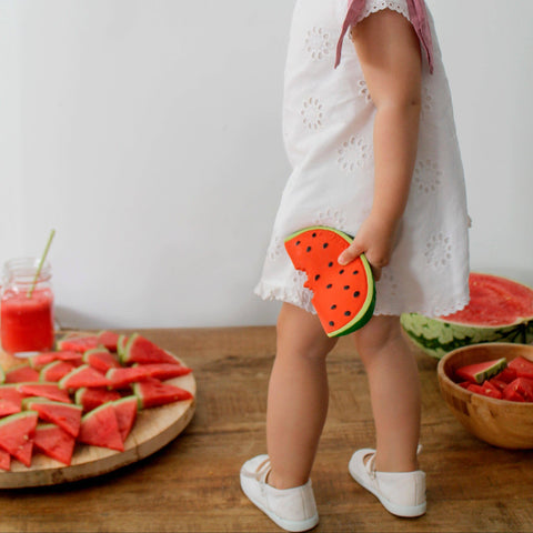 Child in a white dress with watermelon decorations and slices on a wooden table.