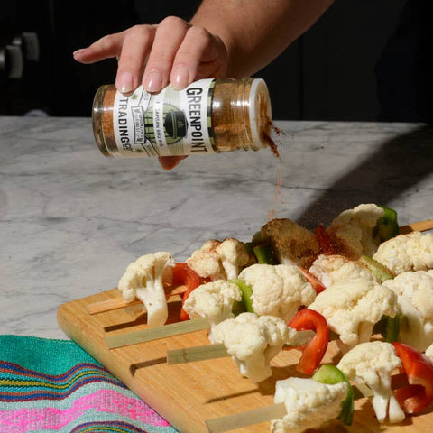 Person seasoning vegetable skewers on a wooden board with a bottle of seasoning.