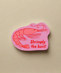 Pink sticker with a shrimp illustration and text 'Shrimply the best!' on a white background