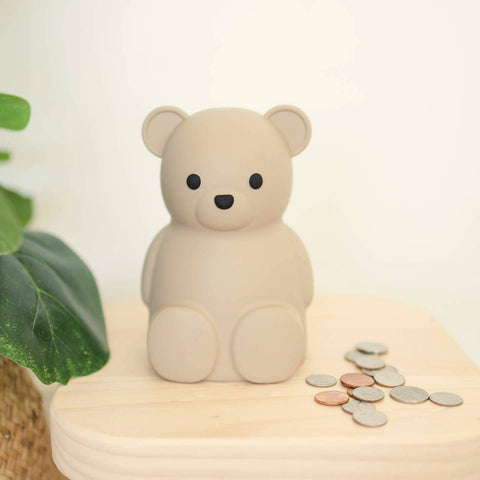 Teddy bear-shaped piggy bank with coins on a wooden base next to a plant