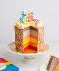 Colorful birthday cake with a slice taken out on a white plate, surrounded by colorful napkins.
