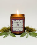 An amber glass jar candle labeled 'Afrevia Ember Dance' sits on a wooden table, with a large beige container in the background.