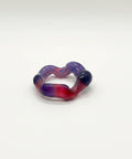 A handmade glass ring with a wavy, heart-like shape in a marbled mix of purple, red, and orange, resting on a light wooden table.