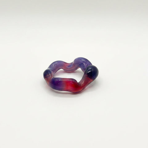 A handmade glass ring with a wavy, heart-like shape in a marbled mix of purple, red, and orange, resting on a light wooden table.