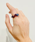 A close-up of a person's hand wearing a twisted ring made of dark purple and blue glass, set against a white background.