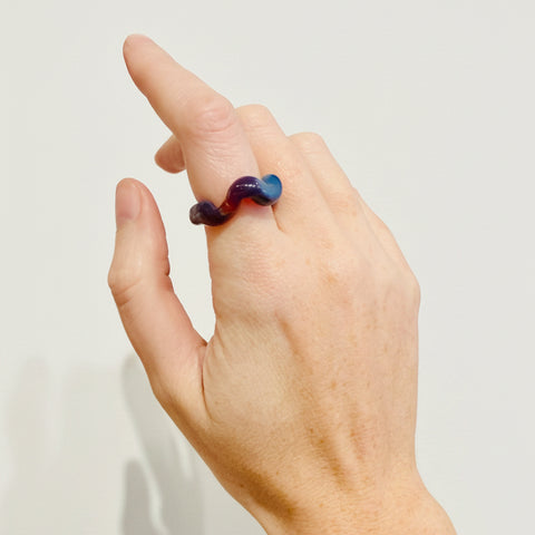 A close-up of a person's hand wearing a twisted ring made of dark purple and blue glass, set against a white background.