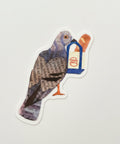 A die-cut sticker featuring a pigeon collage made from book pages, with a small blue and orange bird feeder on the right, placed on a wooden tabletop.