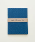 A small blue fabric pouch or notebook cover with a tan paper band reading 'made with denim bits,' placed on a light wood surface.