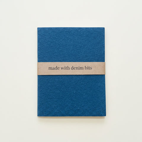 A small blue fabric pouch or notebook cover with a tan paper band reading 'made with denim bits,' placed on a light wood surface.