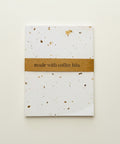 A small white paper packet with scattered coffee bean bits and a brown paper band reading 'made with coffee bits,' placed on a warm wood surface.