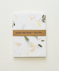 A square handmade paper notebook or journal with a brown paper band reading 'made with floral + fern bits', placed on a light wood surface. The paper has a white base with scattered botanical inclusions in soft pink, yellow, gray, and olive tones.