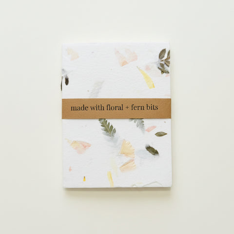 A square handmade paper notebook or journal with a brown paper band reading 'made with floral + fern bits', placed on a light wood surface. The paper has a white base with scattered botanical inclusions in soft pink, yellow, gray, and olive tones.