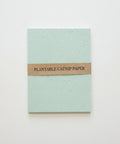 A square sheet of pale mint green plantable catnip paper with a brown paper band around the middle reading 'PLANTABLE CATNIP PAPER', placed on a light wood surface.