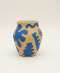 A handmade ceramic pot with a speckled beige and blue pattern, sitting on a wooden table with a laptop blurred in the background.