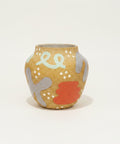 A handmade ceramic pot with a speckled mustard yellow glaze, decorated with light blue abstract loops, gray organic shapes, white dots, and a red-orange splash, sitting on a wooden table with a blurred laptop in the background.