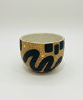 A handmade ceramic tea cup with a speckled beige interior and exterior decorated with black abstract patterns, gold dots, and small yellow accents, sitting on a wooden table with a blurred laptop in the background.