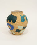 A small handmade ceramic pot with a speckled beige surface, decorated with abstract hand-painted shapes in teal, blue, black, and white, sitting on a wooden table with a laptop in the background.