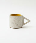 A speckled ceramic mug with a triangular handle sits on a wooden desk in front of a closed gold laptop.