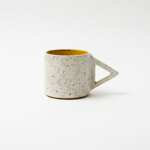 A speckled ceramic mug with a triangular handle sits on a wooden desk in front of a closed gold laptop.
