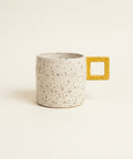 A speckled ceramic mug with a yellow square handle sits on a wooden desk in front of a closed gold laptop.