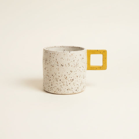 A speckled ceramic mug with a yellow square handle sits on a wooden desk in front of a closed gold laptop.