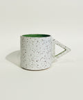 A speckled ceramic mug with a triangular handle, featuring a white exterior with dark brown and black speckles and a green interior, sitting on a wooden surface against a white wall.